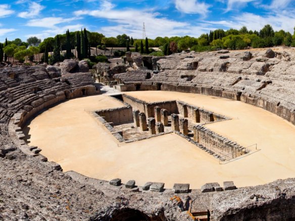 Aerial view of the Itálica amphitheater in Santiponce, Spain, showcasing its large oval structure with tiered seating. Built in the 1st century A.D., it is the largest Roman amphitheater outside Italy, designed for public spectacles such as gladiator fights and animal hunts.